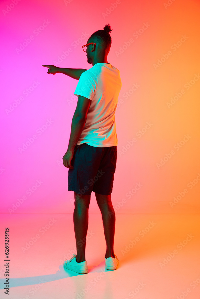 Full length image of backview african man in glasses holding his hand up and pointing something over orange-pink background in neon. Full length image of backview african man in glasses holding his hand up and pointing something over orange-pink background in neon.