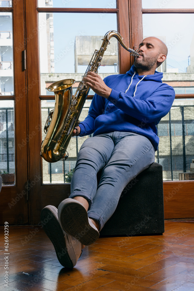 Naklejka premium young latin man sitting at home inspired and excited playing the saxophone.
