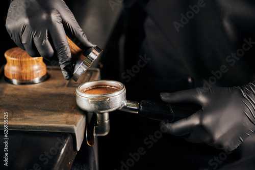 cropped view of barista in black latex gloves pressing ground coffee in portafilter, coffee shop