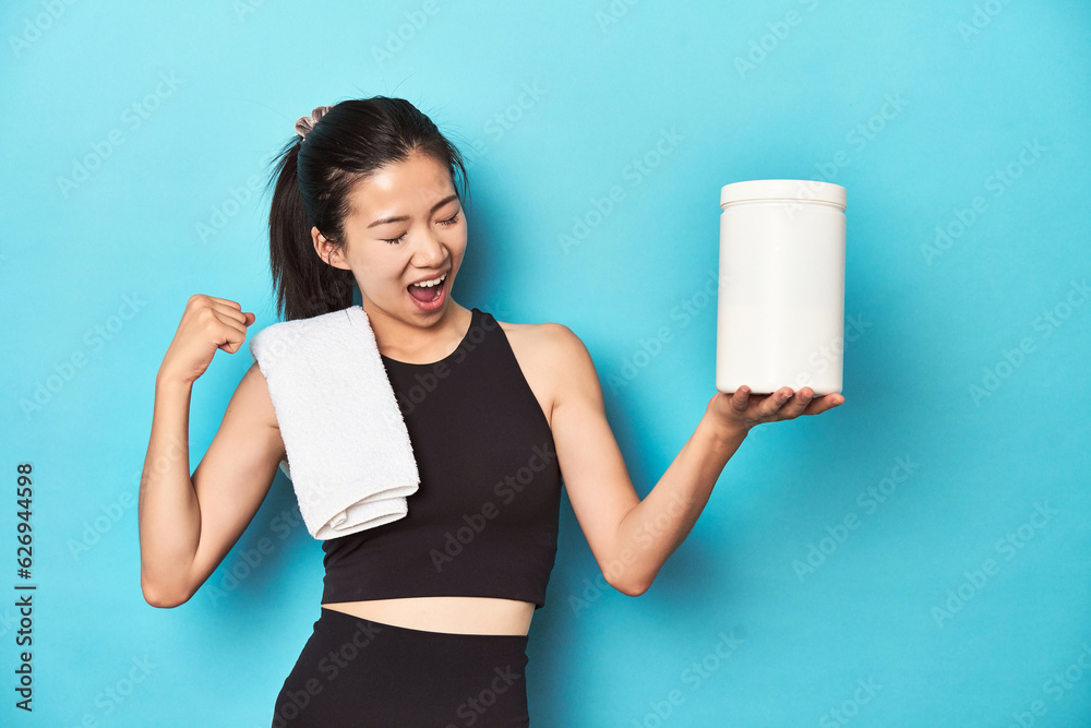 Young Asian sportswoman with protein bottle, gym setup, raising fist after a victory, winner concept. Young Asian sportswoman with protein bottle, gym setup, raising fist after a victory, winner concept.
