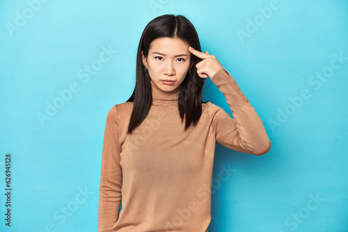 Young Asian woman in brown turtleneck, pointing temple with finger, thinking, focused on a task. © Asier