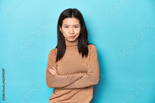Young Asian woman in brown turtleneck, frowning face in displeasure, keeps arms folded.