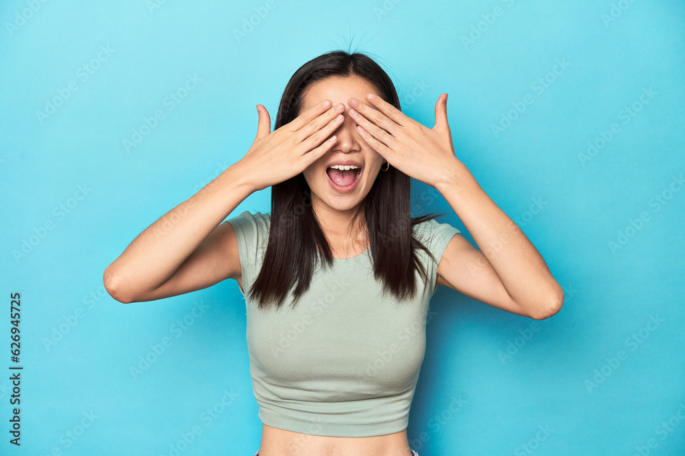 Asian woman in summer green top, studio backdrop, covers eyes with hands, smiles broadly waiting for a surprise.