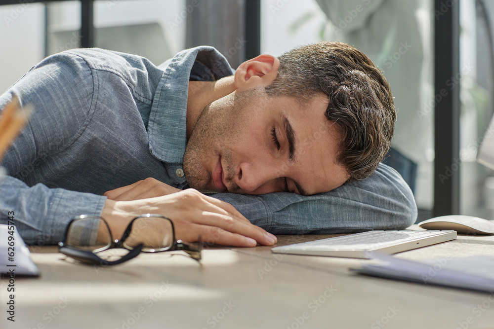Obraz premium Tired businessman sleeping on his desk in front of laptop, overworking, feeling exhausted in modern office.