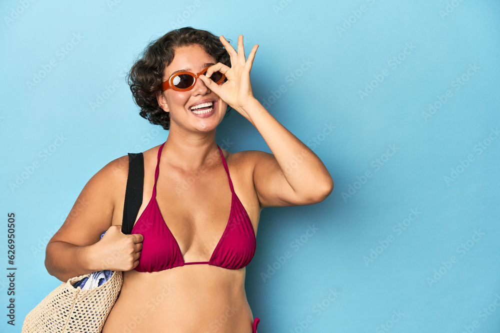 Young woman in bikini with beach bag excited keeping ok gesture on eye.