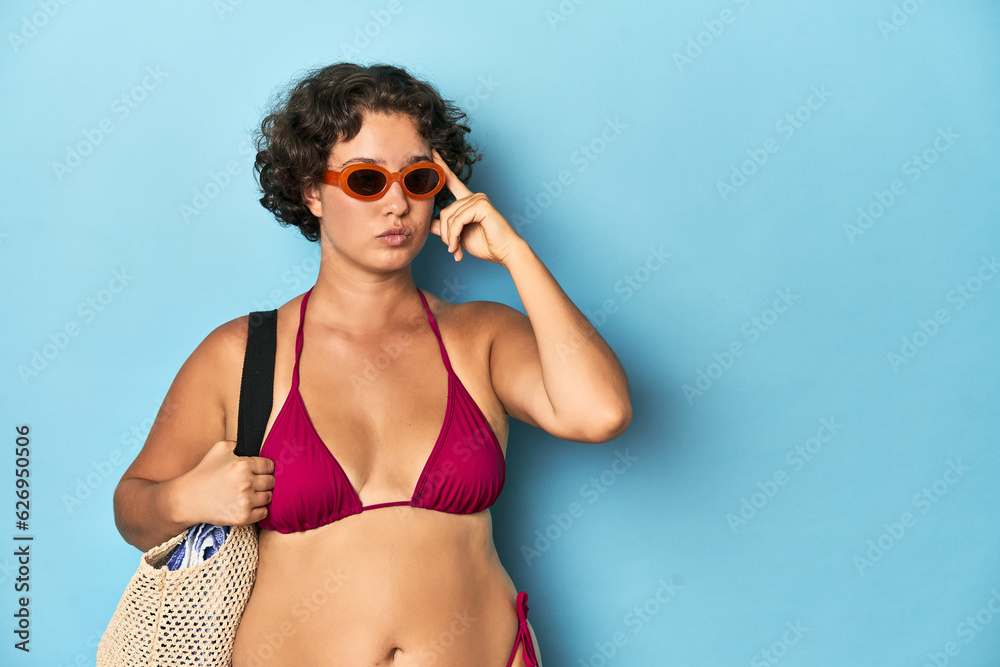 Young woman in bikini with beach bag pointing temple with finger, thinking, focused on a task.