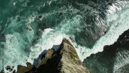 A sharp rock from a bird's eye view, below is a powerful ocean, its waves are breaking against the rock. Intense blue ocean, with huge waves. Drone photo, top to bottom.