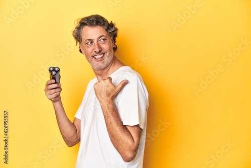 Man holding a razor, yellow studio background points with thumb finger away, laughing and carefree.