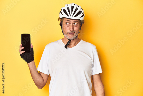 Cyclist man showing phone on yellow backdrop confused, feels doubtful and unsure.