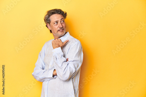 Middle-aged man posing on a yellow backdrop points with thumb finger away, laughing and carefree.