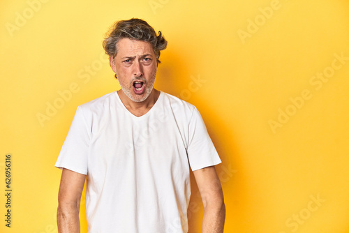 Middle-aged man posing on a yellow backdrop shouting very angry, rage concept, frustrated. © Asier