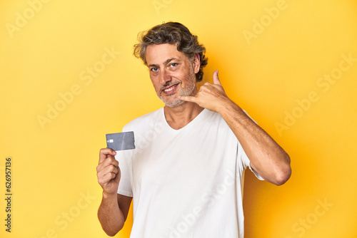 Man holding a credit card, yellow studio backdrop showing a mobile phone call gesture with fingers.