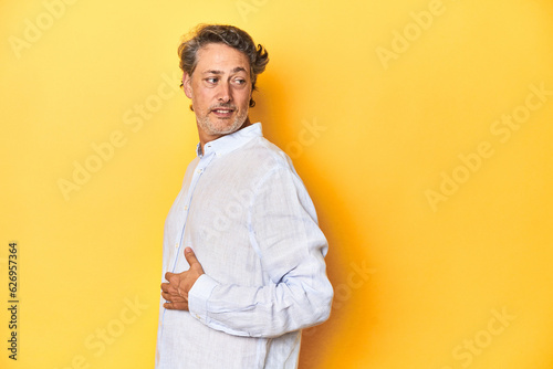 Middle-aged man posing on a yellow backdrop looks aside smiling, cheerful and pleasant.