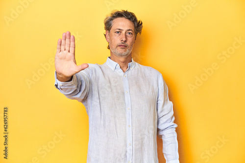 Middle-aged man posing on a yellow backdrop standing with outstretched hand showing stop sign, preventing you.