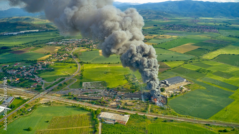 Fototapeta premium A black column of smoke and a fire in a warehouse, against the backdrop of the city. Photo from above from a drone.