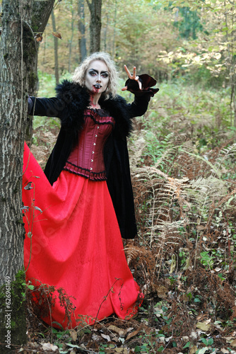 A woman in the image of a vampire or a witch with white make-up and in a medieval dress poses in the forest in front of the camera