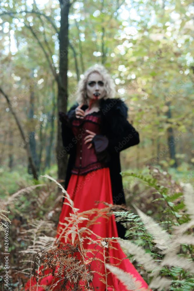 A woman in the form of a vampire or witch in a coat, corset and long red skirt. Blood trickles down the vampire's chin. Focus on plants A woman in the form of a vampire or witch in a coat, corset and long red skirt. Blood trickles down the vampire's chin. Focus on plants
