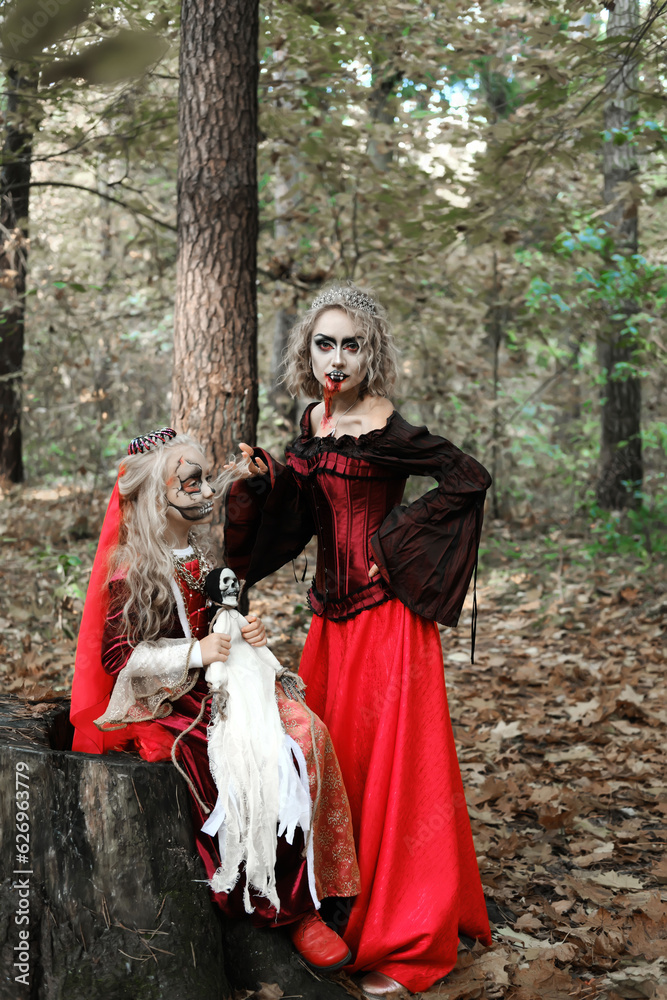 A woman in the form of a vampire or a sorceress and a girl in the form of a dead princess with a skeleton doll in their hands pose in the forest. Models are looking at camera
