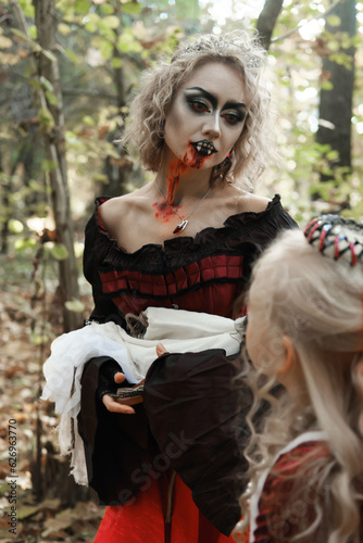 A woman in the form of a vampire or a sorceress holds a doll in the form of a skeleton in her arms and looks at a princess girl in fairy-tale makeup and medieval dress. Image for Halloween. 