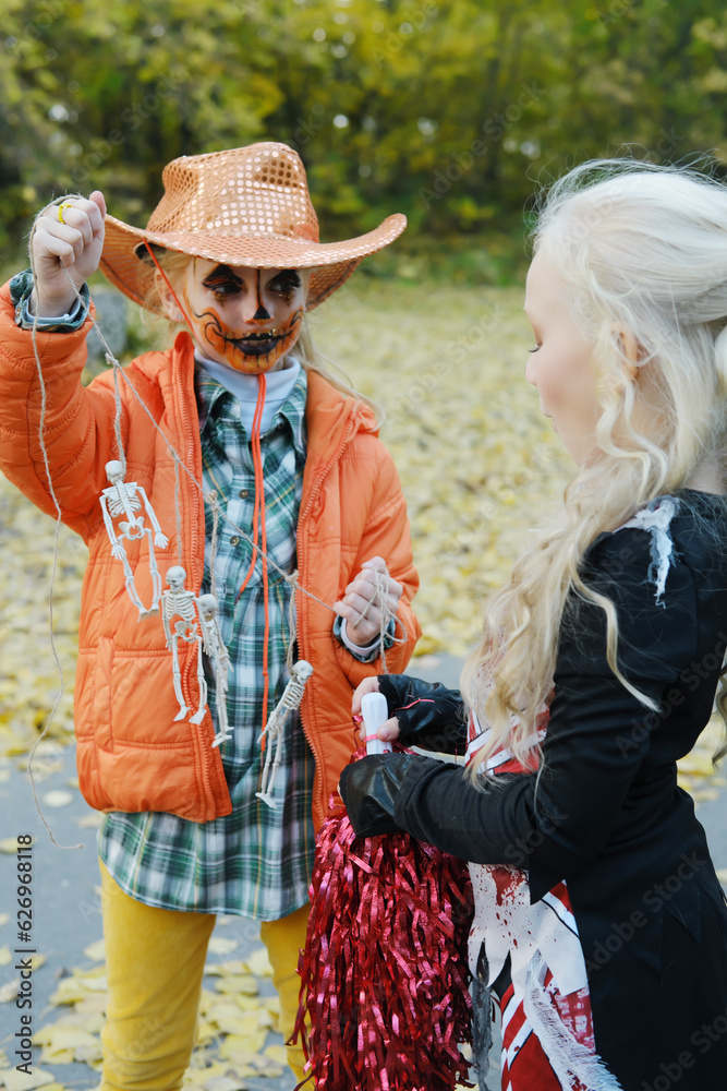 Girl child in an orange jacket and a cowboy hat shows her friend in a cheerleader costume her skeletons on a rope. Halloween celebration