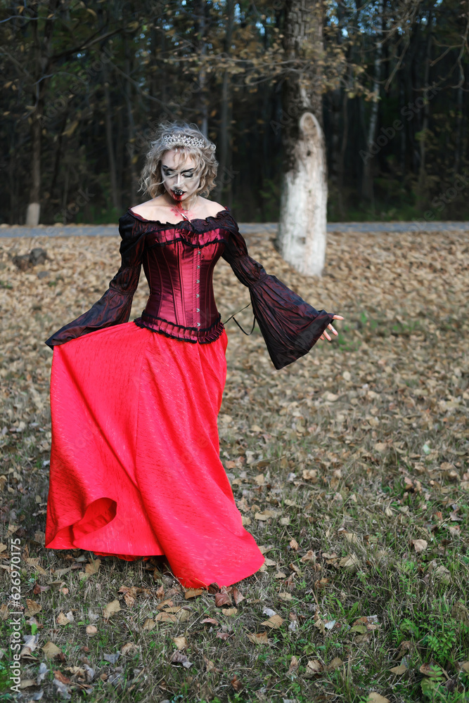 A woman in the form of a vampire or a witch in a corset and a long red skirt. Blood trickles down the vampire's chin. Woman walking in the field