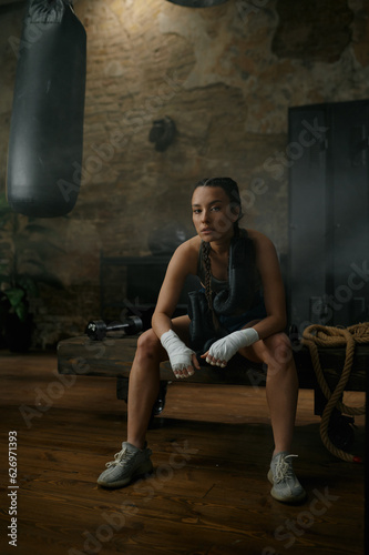 Female boxer in boxing bandage carrying gloves on shoulders sitting on bench