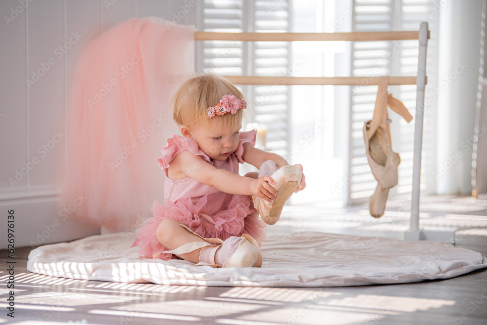 Fototapeta premium A cute little ballerina in a pink ballet costume sits near the barre in the room and tries to put on her pointe shoes. Kid and ballet dance. Copyspace.