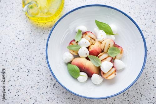 Plate of salad with grilled peaches, mini mozzarella and green basil, horizontal shot on a light-grey granite background, elevated view