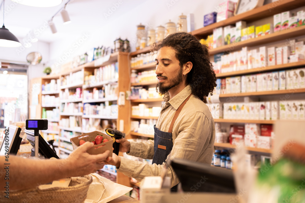 Young ethnic male cashier scanning products during work in eco friendly store
