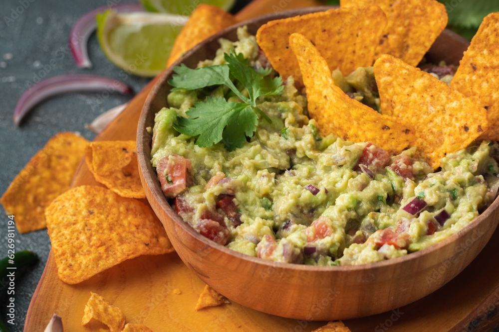 Wooden bowl of traditional Mexican guacamole with nachos on grey concrete background. Tortilla chips with guacamole sauce dip and ingredients: avocado, cilantro, onion, hot pepper and lime