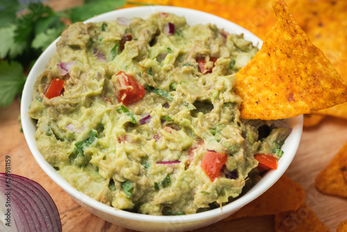 White bowl of traditional Mexican guacamole with nachos on grey concrete background. Tortilla chips with guacamole sauce dip and ingredients: avocado, cilantro, onion, hot pepper and lime