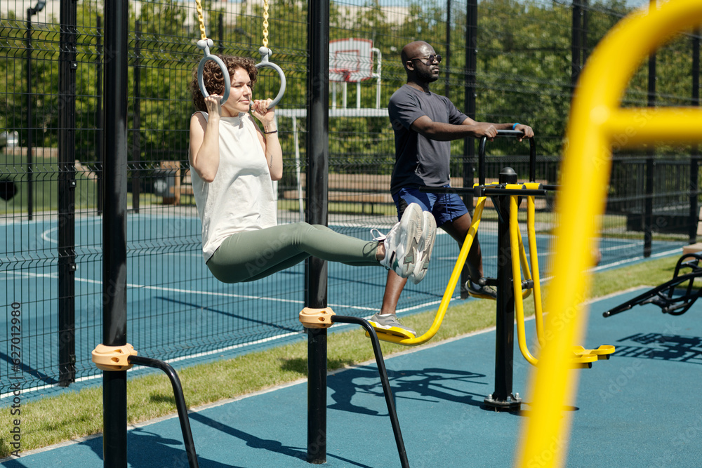 Young brunette sportswoman hanging on still rings and doing L sits on sports ground against African American man exercising on facilities