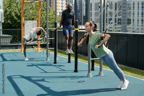 Young pretty and fit woman in activewear hanging on still rings on sports ground while doing physical exercises against intercultural people