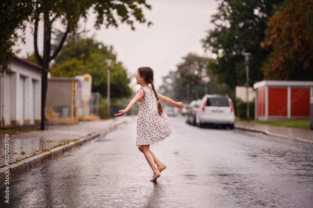 Little preschooler girl in white dress with black hearts dancing and spinning on wet empty street under the rain