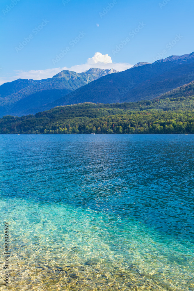 A vertical view of a lake with clear blue-green waters, some alpine mountains at background and the moon over a blue sky, in a summer day. Refreshing and relaxing scene. Lake Bohinj, Slovenia.