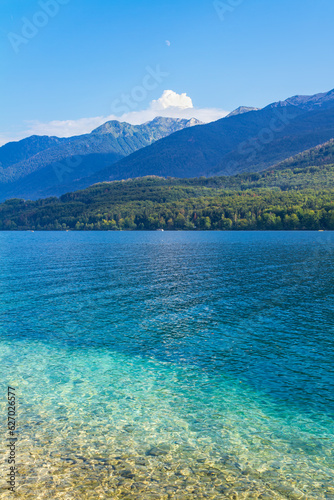 A vertical view of a lake with clear blue-green waters, some alpine mountains at background and the moon over a blue sky, in a summer day. Refreshing and relaxing scene. Lake Bohinj, Slovenia.