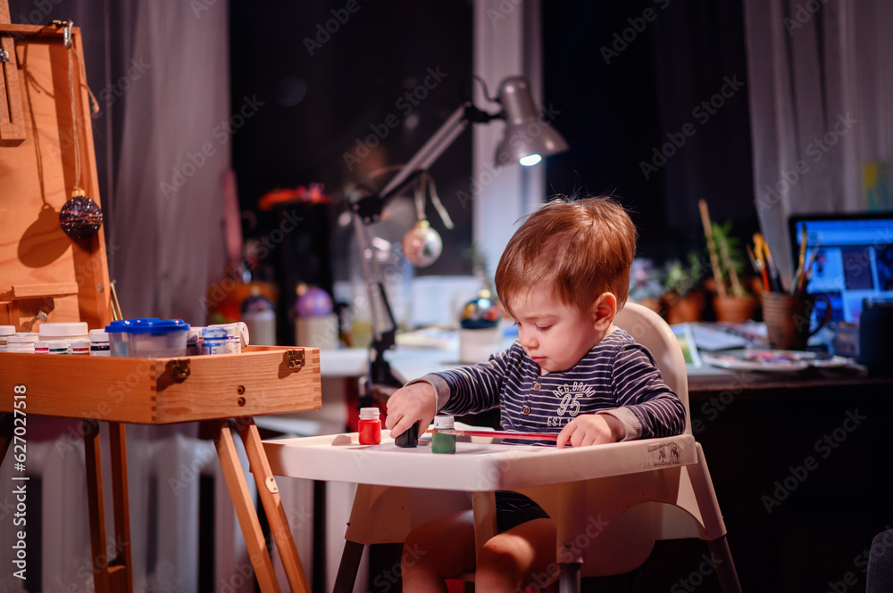 Lottle toddler boy sitting in chair for babies happy to play with brush and jars of paint at home Lottle toddler boy sitting in chair for babies happy to play with brush and jars of paint at home