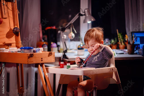 Lottle toddler boy sitting in chair for babies happy to play with brush and jars of paint at home