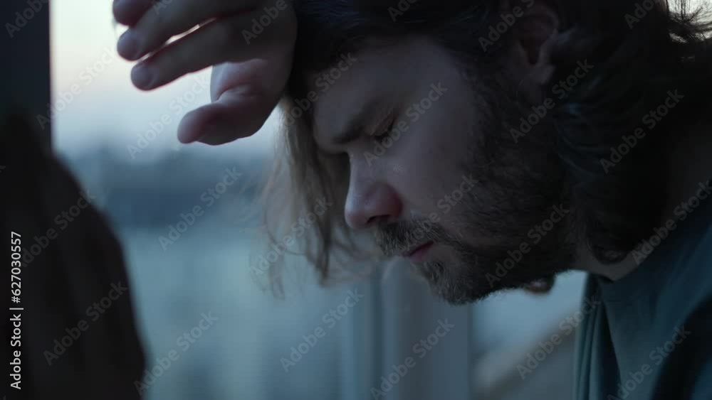 Depressed man close eyes, thinking about problems near window. Close up of pensive, sad depressed unhappy beard man with long hair. Mental health, man in tears, man cry depression concept. Authentic