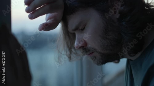 Depressed man close eyes, thinking about problems near window. Close up of pensive, sad depressed unhappy beard man with long hair. Mental health, man in tears, man cry depression concept. Authentic