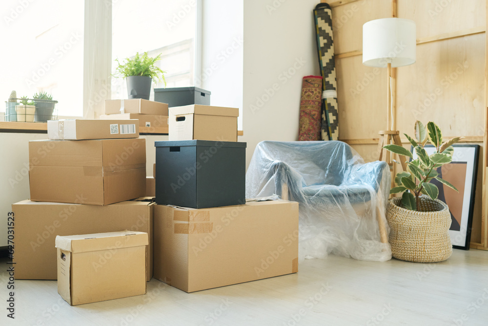 Stacks of large boxes standing against windowsill with green domestic plants in flowerpots and corner with rolled carpets and armchair Stacks of large boxes standing against windowsill with green domestic plants in flowerpots and corner with rolled carpets and armchair