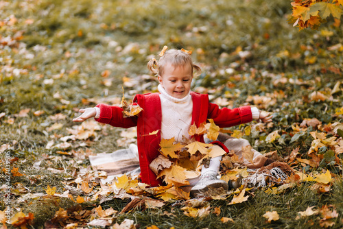 A little girl in a red warm coat is playing with yellow leaves  having fun and smiling sitting on a plaid plaid in an autumn park outdoors in fall. Selective focus