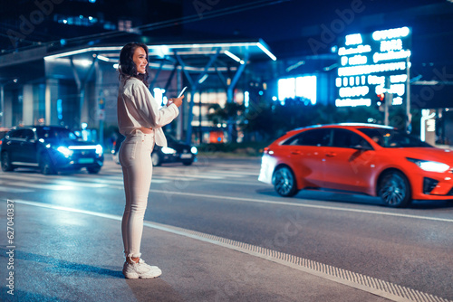 Full length shot of young woman standing on the street in the night holding smartphone, waiting taxi, free space © Prostock-studio