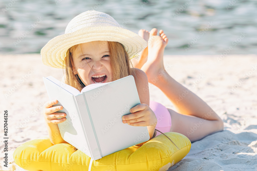 Happy cute little blonde girl in straw hat reading book at beach. Beach vacation concept