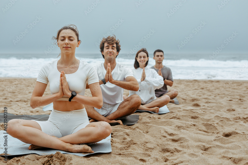 The beachside lotus. Young people meditating together during yoga lesson outdoors, sitting on mats on the beach The beachside lotus. Young people meditating together during yoga lesson outdoors, sitting on mats on the beach