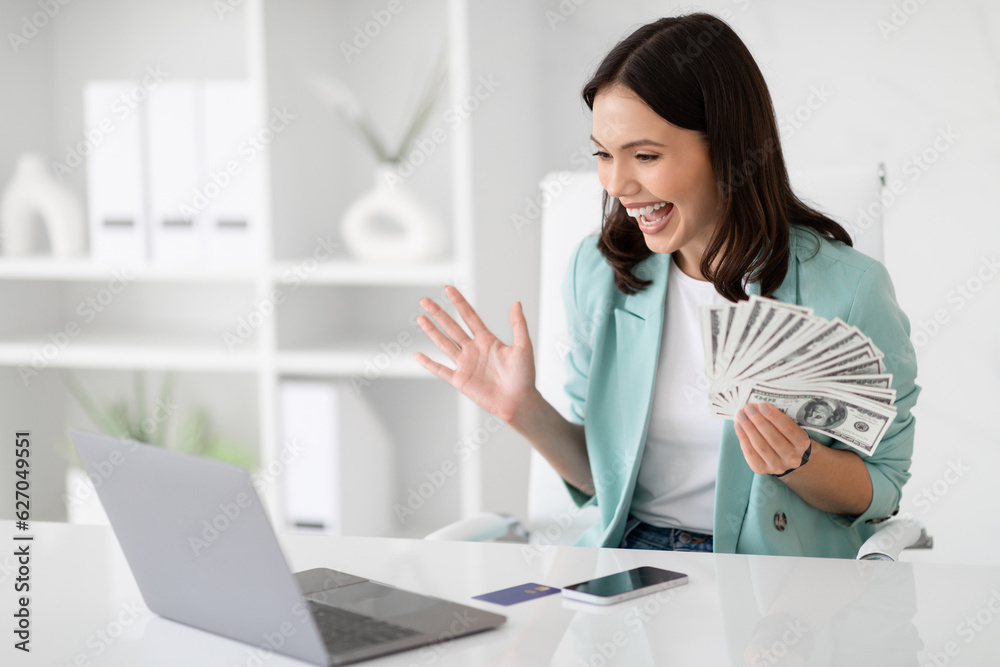 Surprised glad millennial caucasian lady in suit hold lot of cash, enjoy success, celebrate win, look at laptop Surprised glad millennial caucasian lady in suit hold lot of cash, enjoy success, celebrate win, look at laptop