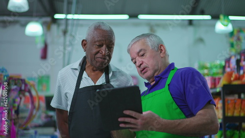 Diverse Senior Employees at Supermarket holding tablet device pointing at Grocery Store inventory, Teamwork Scene of Older Manager Guiding Employee