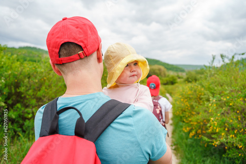 A father with a backpack holds a baby toddler around his neck  hiking in the mountains in summer