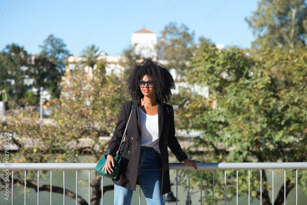 Young beautiful black woman with afro hair dressed in casual clothes and leaning on the railing while enjoying her holidays in seville, spain. In the background the river and the flamenco quarter.