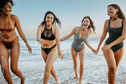 Happy young diverse women having fun on the beach, running in water and laughing, enjoying summer time together